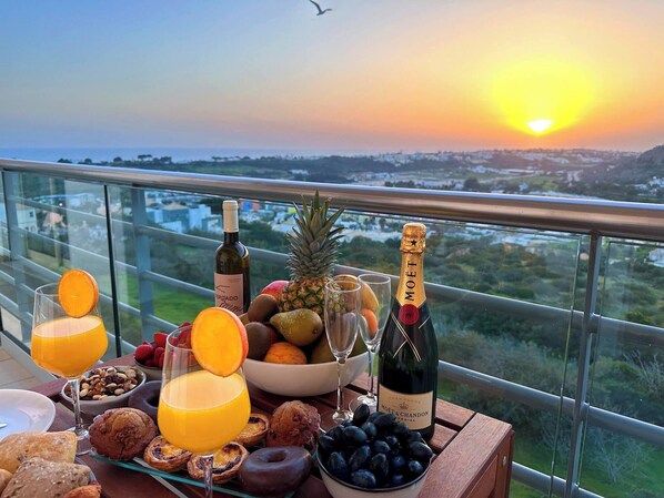 A table with food, drinks, and a bottle of champagne on a balcony overlooking a landscape.