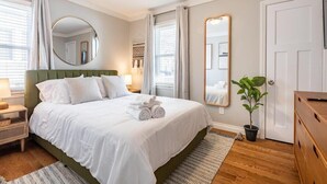 Ein ordentlich eingerichtetes Schlafzimmer mit einem großen Bett, einem Nachttisch mit Lampe, einem Spiegel und einem Blumentopf.