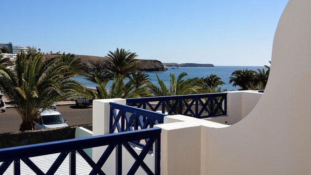 A balcony with a view of the sea, palm trees, and a building in the background.