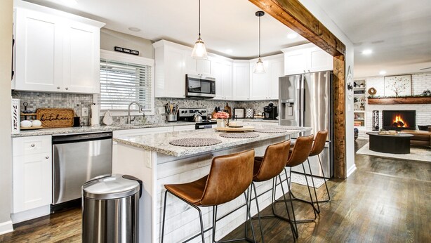 Una cocina moderna con armarios blancos, una isla central con taburetes de bar, electrodomésticos de acero inoxidable y una chimenea en la zona de estar.
