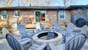 Un patio con una fogata circular rodeada de sillas Adirondack, una terraza de madera y una casa de ladrillo con una ventana que da al deck.