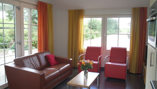 A modern living room with a brown leather sofa, a wooden coffee table, and two red chairs. There are large windows with yellow curtains, and a view of greenery outside.
