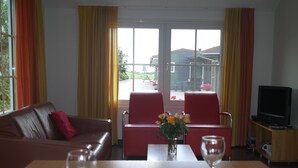 Un salon avec un canapé en cuir marron, des fauteuils rouges, une table à manger ornée d’un vase de fleurs, un téléviseur et une fenêtre avec des rideaux.