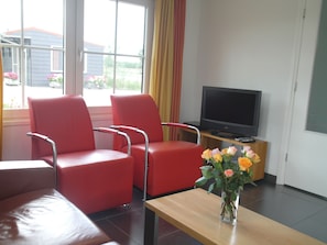 A room with red chairs, a wooden table, a TV, and a vase with flowers.