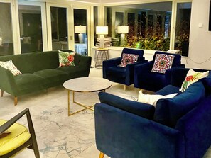 A living room with a green sofa, two blue armchairs, a small round table, and a glass door leading outside.