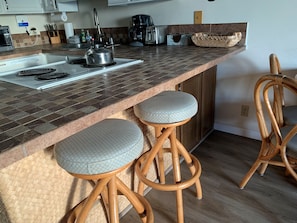 A kitchen with a tiled countertop, a kettle, a coffee maker, and two wooden bar stools with cushioned seats.