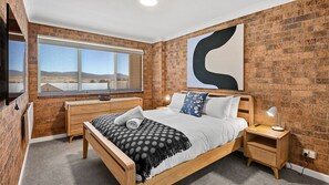 A bedroom with a wooden bed, a large window, a brick wall, and a modern abstract painting.
