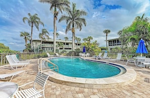 A swimming pool with lounge chairs and umbrellas, surrounded by palm trees and a building with balconies.