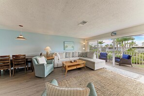A living room with a sofa, coffee table, and chairs, a dining area with a table and chairs, and a large sliding glass door leading to an outdoor area with chairs and a view of palm trees.