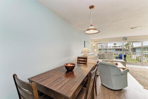 A dining area with a wooden table and chairs, a hanging light fixture, and a view of a building through large windows. - West Bay Cove 226 · Kayak Launch · For Sale (Holmes Beach)