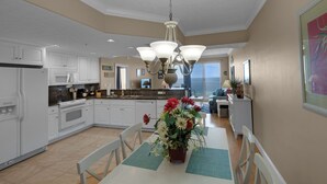 A spacious kitchen with white cabinets, a granite countertop, and a central island. There is a dining area with a table and chairs, and a large window offering a view of the ocean.