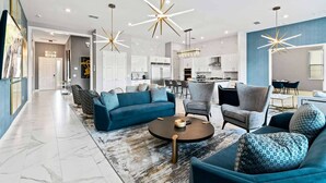 Una sala moderna con un sofá modular, sillones y una mesita de centro. Se aprecia una cocina al fondo con armarios blancos y una mesa de comedor.