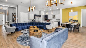 Un salon moderne avec un canapé d’angle, un coin repas doté d’une longue table et une cuisine avec des meubles blancs et un mur d’accent jaune.