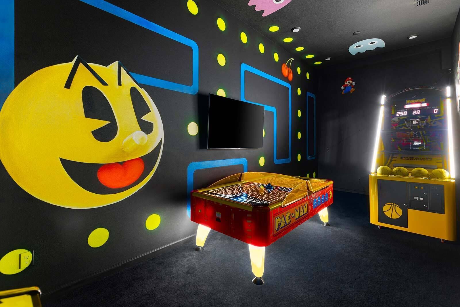 A room with a Pac-Man arcade machine, a large Pac-Man mural, and a television screen.