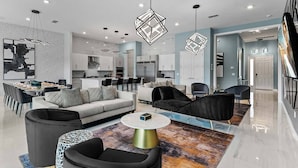 Ein modernes Wohnzimmer mit einer Couch, Sesseln, einem Couchtisch und einem Teppich. Im Hintergrund befindet sich eine Küche mit weißen Schränken und ein Essbereich mit Tisch und Stühlen.