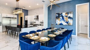 A modern kitchen with white cabinets, a stainless steel refrigerator, and a dining area with a glass table and blue chairs. - Feature-Rich Home w Superhero Theme BR, Huge Arcade & Theater - Near Disney (Davenport)