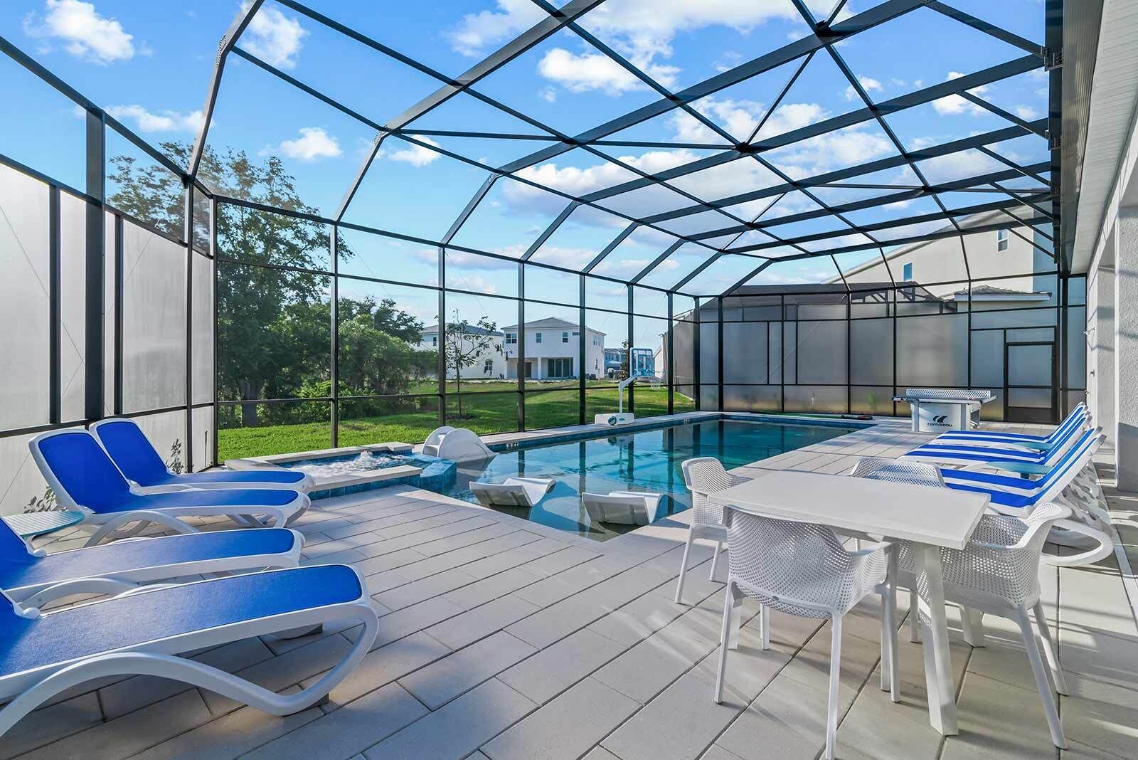 Zona de piscina exterior cubierta con sillones reclinables y una mesa, rodeada por una estructura metálica y paneles de vidrio.