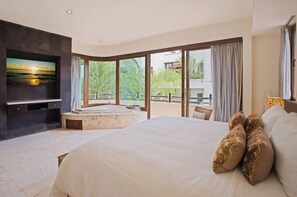 A spacious bedroom with a large bed, a built-in TV, a jacuzzi, and a balcony with a view of greenery.