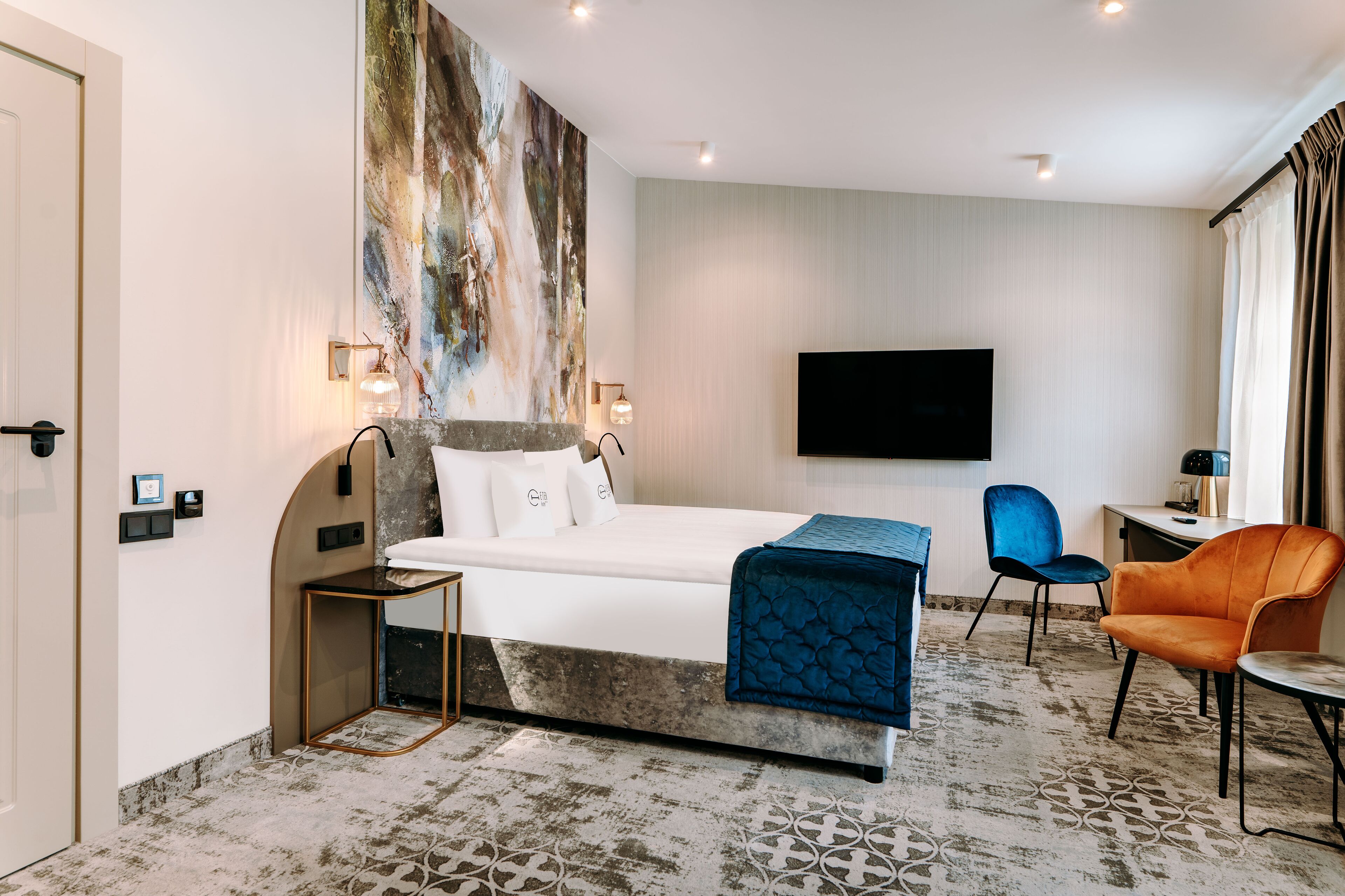 Ein Hotelzimmer mit einem Bett, einem Schreibtisch, einem Stuhl, einem Fernseher und einem großen, abstrakten Gemälde an der Wand.