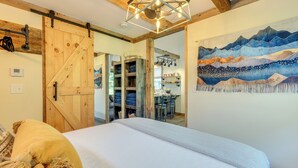 Ein Schlafzimmer mit einem großen Bett, Holzbalkendecke, einer Schiebetür im Bauernhausstil, einer Küchenzeile mit Esstisch und einem Wandbild mit Berglandschaft.