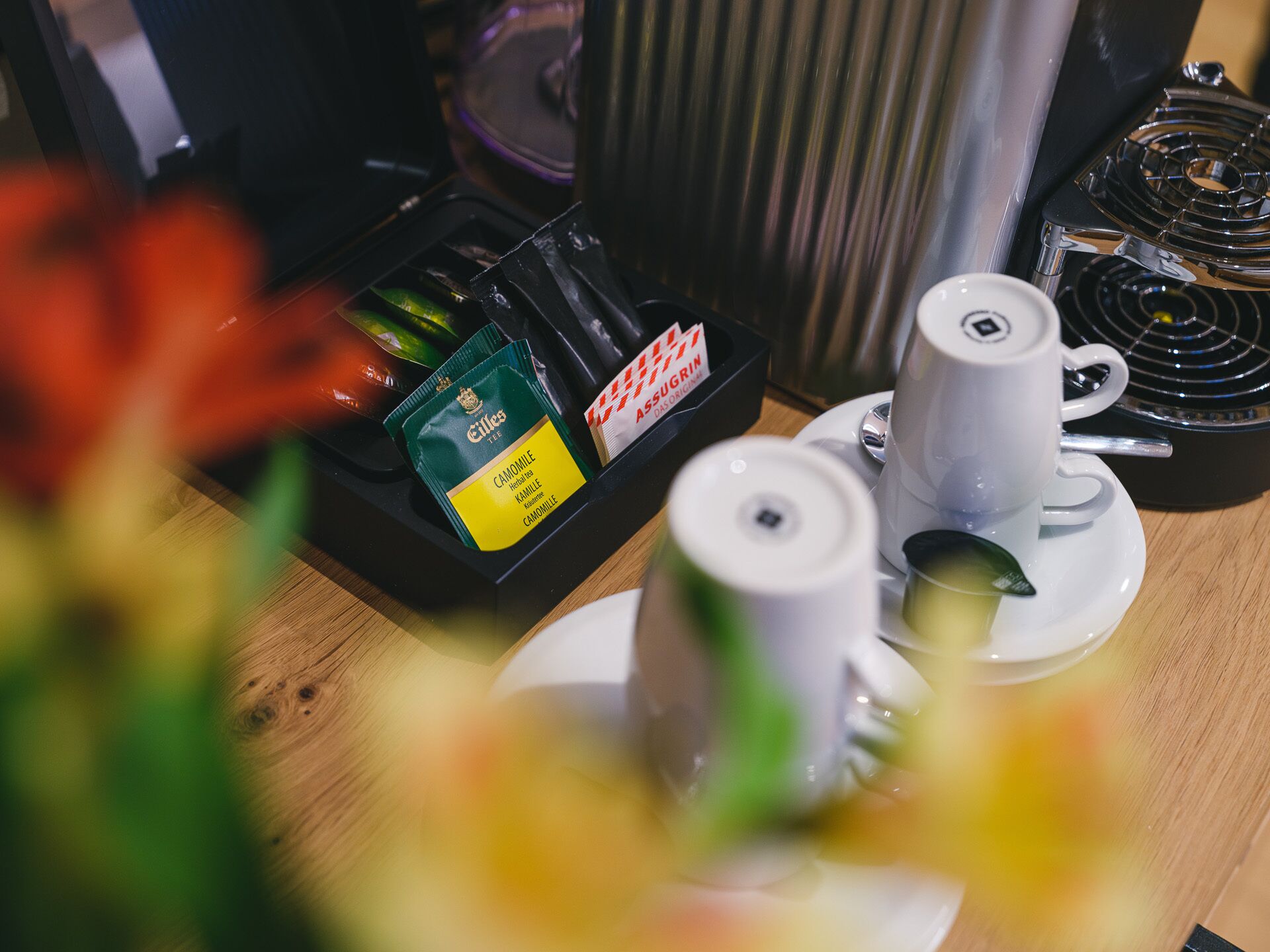 Een koffiehoek met twee witte kopjes op schoteltjes, een koffieapparaat en een dienblad met theezakjes en een doosje met 'Assugrin'.