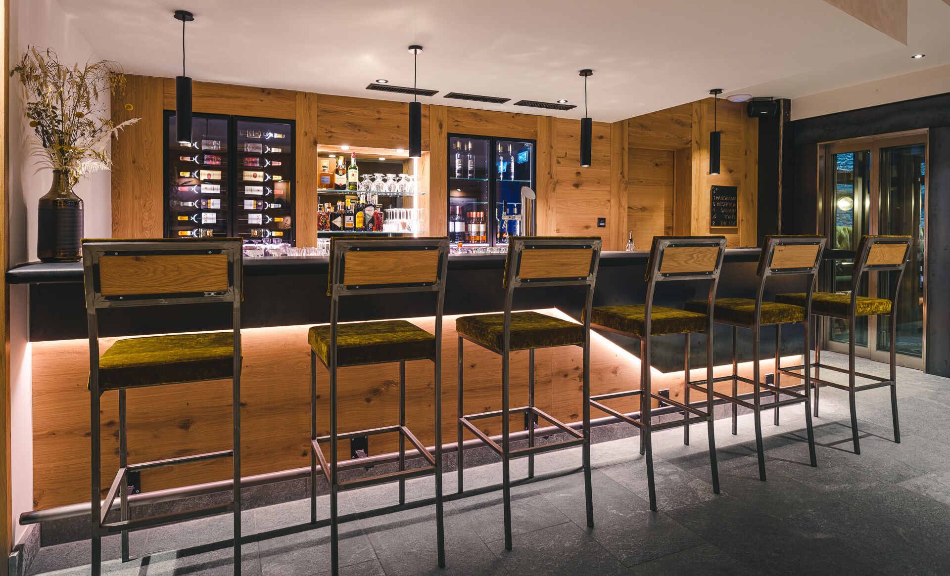 Una zona de bar con taburetes de madera, una vitrina de vidrio con botellas y una pizarra de menú.