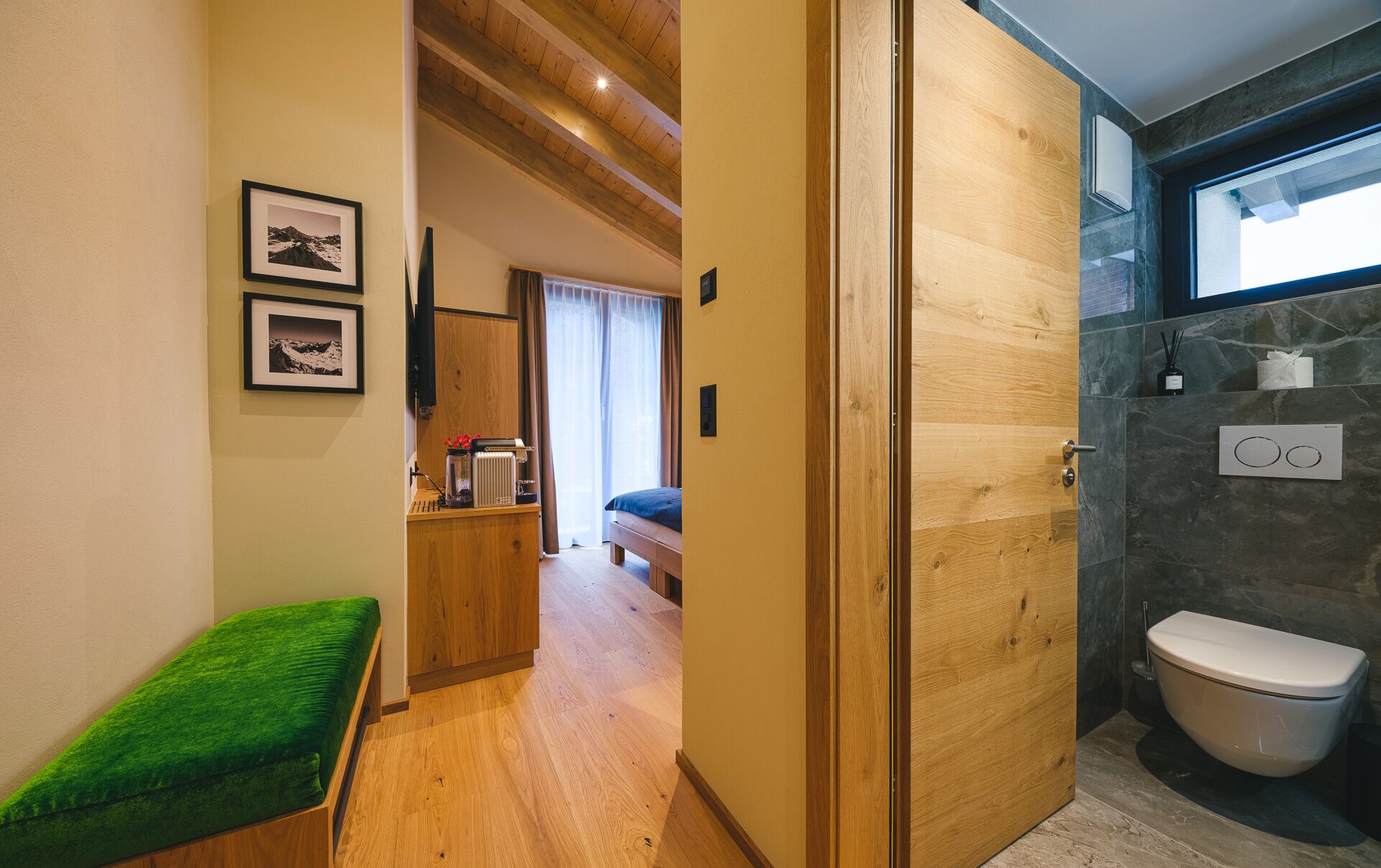 Habitación de hotel con un banco de madera, una cama, un baño con inodoro y ducha, y un pasillo que conduce a otra habitación.