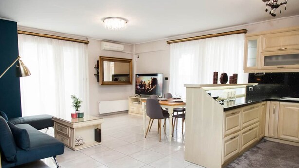 A modern living room with a sofa, a dining area with a table and chairs, a kitchen with cabinets and a countertop, and a television. - Elegant & Spacious  Apt. with Sea View (Piraeus)