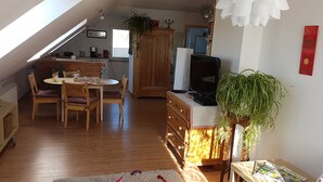 Une pièce chaleureuse et bien éclairée, avec une table et des chaises en bois, un téléviseur et une grande fenêtre laissant entrer la lumière naturelle.