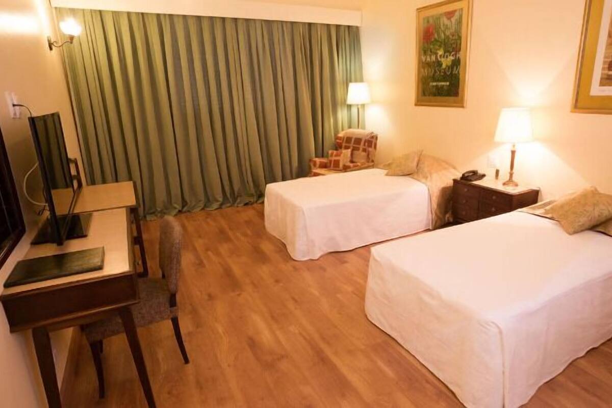 Hotellihuone, jossa on kaksi erillistä sänkyä, työpöytä tuolilla, lamppu ja seinällä oleva kehystetty kuva.