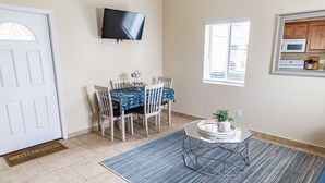 A dining area with a table and chairs, a television mounted on the wall, a window with blinds, and a kitchen area with cabinets and a microwave. - The PB Spot 1BR w/ Parking (San Diego)