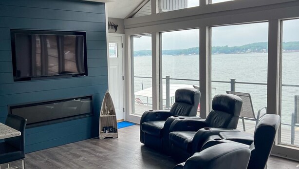 Een moderne woonkamer met een groot raam met uitzicht op een waterpartij, een flatscreen televisie aan de muur en een zithoek met lederen fauteuils.