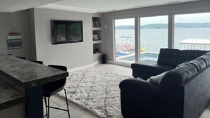 Een moderne woonkamer met een groot raam, een flatscreentelevisie, een donkergrijze bank en een keuken met een marmeren aanrecht.