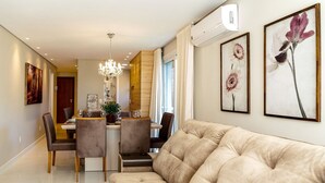 A modern living room with a beige sofa, dining table, and chairs. There is a chandelier, artwork on the walls, and an air conditioning unit. - LOCAR-IN GRAMADO- HOLLYWOOD (Gramado)