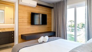 A modern hotel room with a large bed, a flat-screen TV mounted on a wooden wall, a built-in bench, and a large window with curtains. - LOCAR-IN GRAMADO- HOLLYWOOD (Gramado)