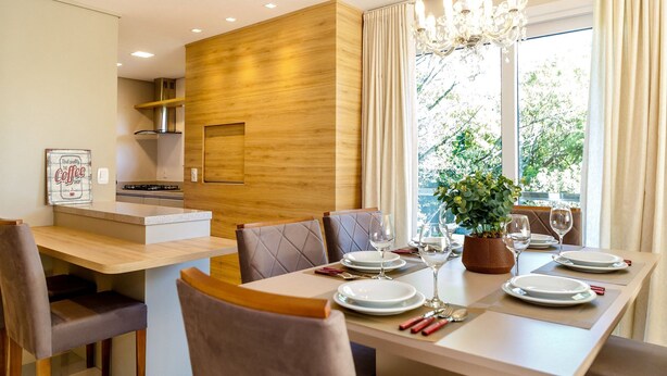 A modern dining area with a wooden accent wall, a chandelier, and a view of trees through large windows. - LOCAR-IN GRAMADO- HOLLYWOOD (Gramado)