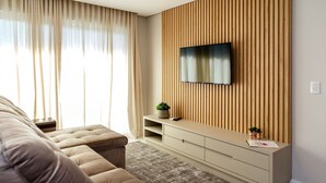 A modern living room with a beige sofa, a flat-screen TV mounted on a wooden panel wall, a small side table with a plant, and a patterned rug. - LOCAR-IN GRAMADO- HOLLYWOOD (Gramado)
