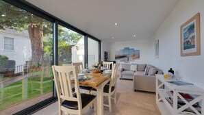 A spacious living area with a dining table, chairs, a sofa, and a bookshelf. There is a large window offering a view of a tree and a house. - 2 Bed in Dartmouth (oc-bc42ab) (Dartmouth)
