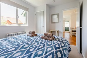 A bedroom with a patterned bedspread, two rolled towels, a radiator, and a view of the outdoors.