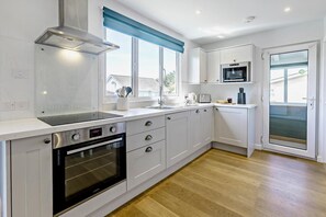 A modern kitchen with a built-in oven, a range hood, and a window with blue blinds. - 2 Bed in Croyde (oc-18lead) (Croyde)