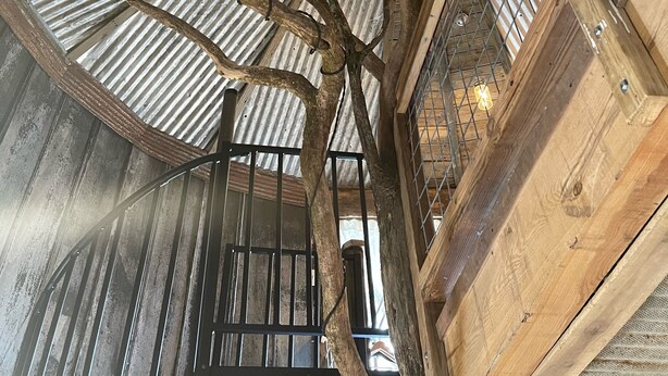 Ein rustikaler Innenraum mit Wellmetall-Decke, Holzbalken und einer zentralen Holzkonstruktion, die an einen Baum erinnert.