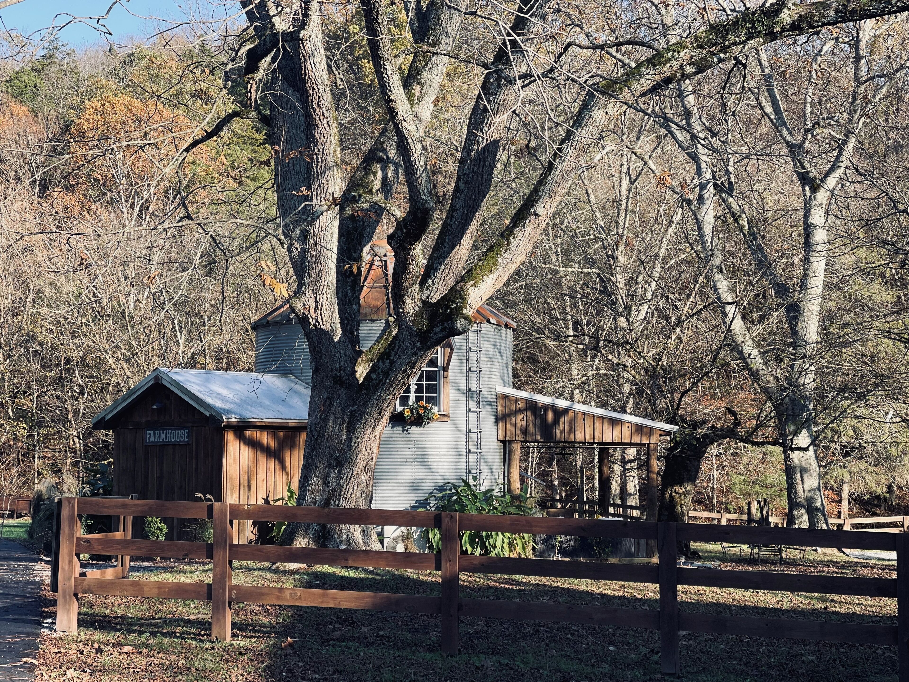 A rustic farmhouse with a large tree in the foreground, a wooden fence, and a metal silo in the background.