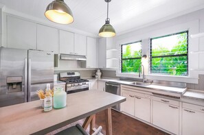 A modern kitchen with stainless steel appliances, white cabinetry, and a large window with a view of greenery. - Lush Poipu Oasis, 4 Bed 4 Bath With Hot Tub, Beach Walk, A/C, Sunsets, & Surf (Koloa)