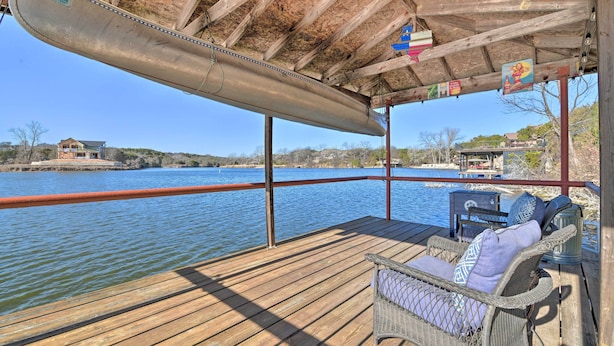 A wooden deck with a view of a lake, a covered area with a metal boat, and a wicker chair with cushions. - Lakefront Getaway w/ Boat Dock, Canoe, Grill! (Granbury)