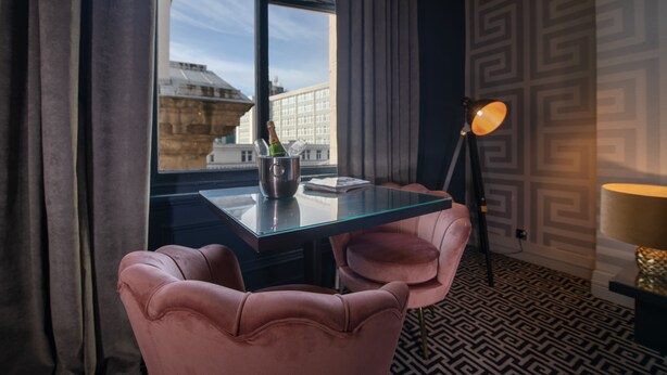 Ett hotellrum med ett glasbord, två rosa stolar, en flaska champagne, en lampa och tapeter med mönster.