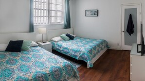 Ein Schlafzimmer mit zwei Betten, einem Fenster mit Jalousien, einem Nachttisch mit Lampe und einer Tür, an der ein Handtuch hängt.