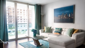 Ein modernes Wohnzimmer mit Glastür zum Balkon, ein weißes Sofa, ein Couchtisch und ein Stadtbild an der Wand.