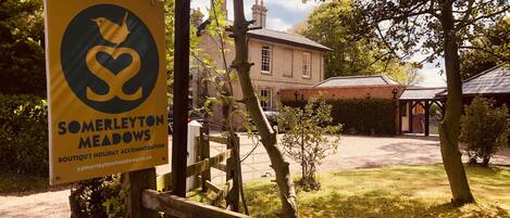 A sign for SOMERLEYTON MEADOWS holiday accommodation with a view of a traditional house and a covered outdoor area.