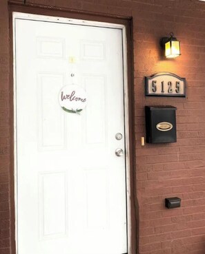 Una puerta blanca con un cartel que dice 'Welcome', un buzón y un letrero con un número de calle en una pared de ladrillo.