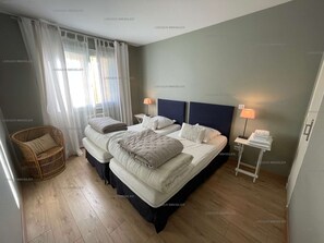 Une chambre avec deux lits, une chaise en osier, une table de chevet avec une lampe et une fenêtre ornée de rideaux légers.